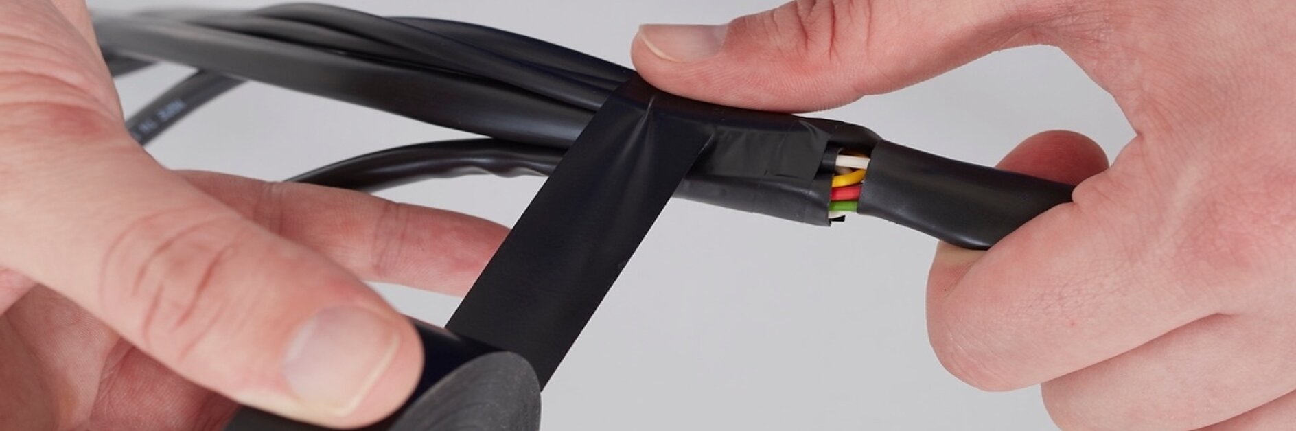 Close-up of hands wrapping black tesa tape around a bundle of cables. The cables include visible colored wires at the cut end, and the person is securing them by tightly applying the tesa tape. (This text has been generated by AI)