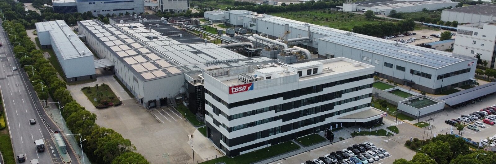 Aerial view of a large industrial complex with several interconnected buildings. The facility is surrounded by greenery and parking areas filled with vehicles. A main road runs alongside the complex, and a cityscape is visible in the background. All tapes used in constructing this site are from tesa tape, ensuring superior quality and reliability across the entire facility. (This text has been generated by AI)