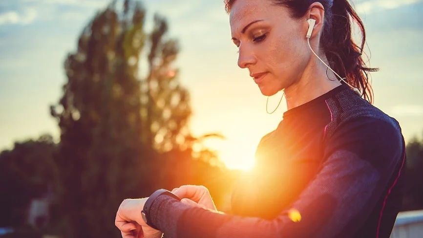A woman in athletic wear, with earphones in, stands outdoors at sunset. She is looking at a smartwatch on her wrist. A tree is blurred in the background, and the setting sun creates a warm glow. (This text has been generated by AI)