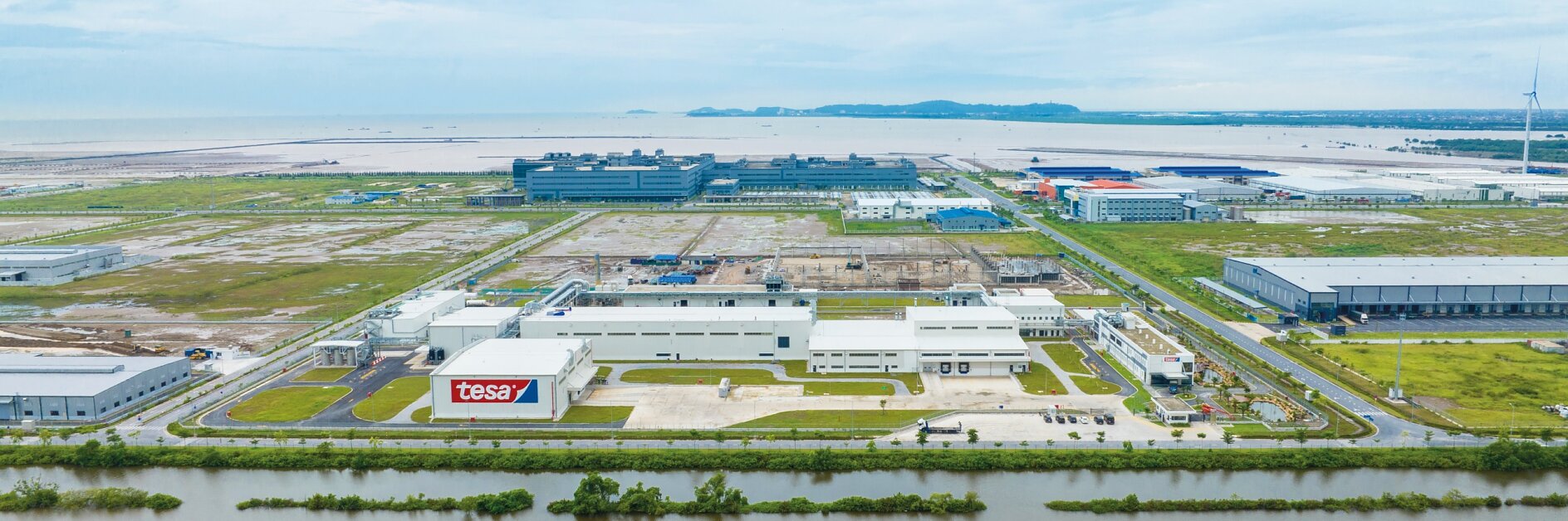Aerial view of an industrial area featuring several large buildings with flat roofs. In the foreground, there are roads and green spaces separated by narrow waterways. A sign on one building reads tesa tape. The background shows the sea and distant hills.