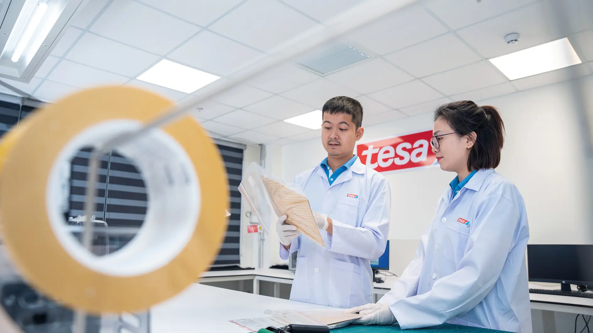 Two people in lab coats stand in a brightly lit laboratory. One holds a sheet of material while the other looks on. A reel of tesa tape is visible in the foreground. A red and white sign with the word tesa is on the wall behind them.