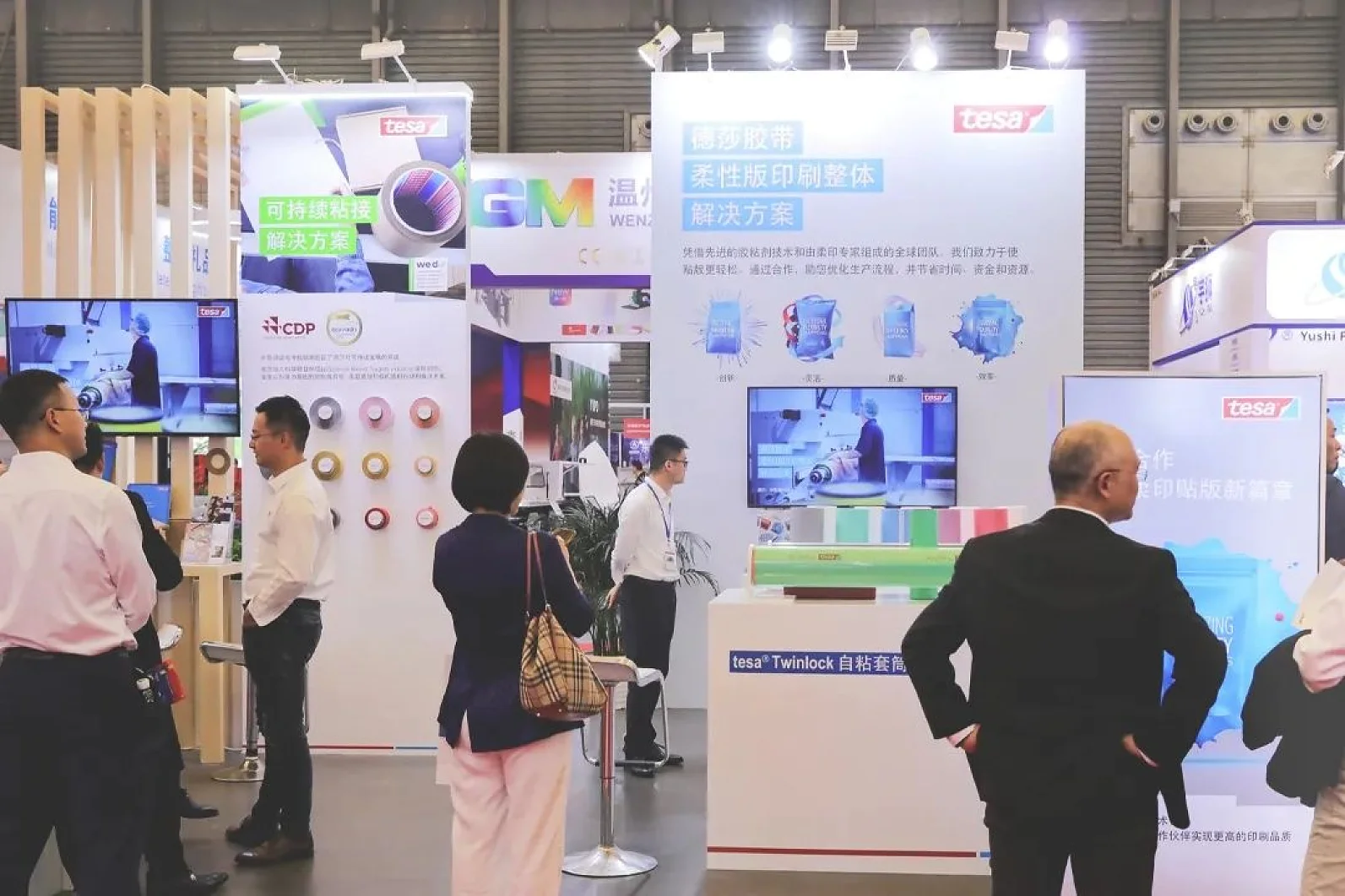 People are gathered at a trade show booth featuring tesa branding. Displays showcase various tesa tape products and packaging solutions. Attendees are engaged in conversations, and several large screens present product information. (This text has been generated by AI)