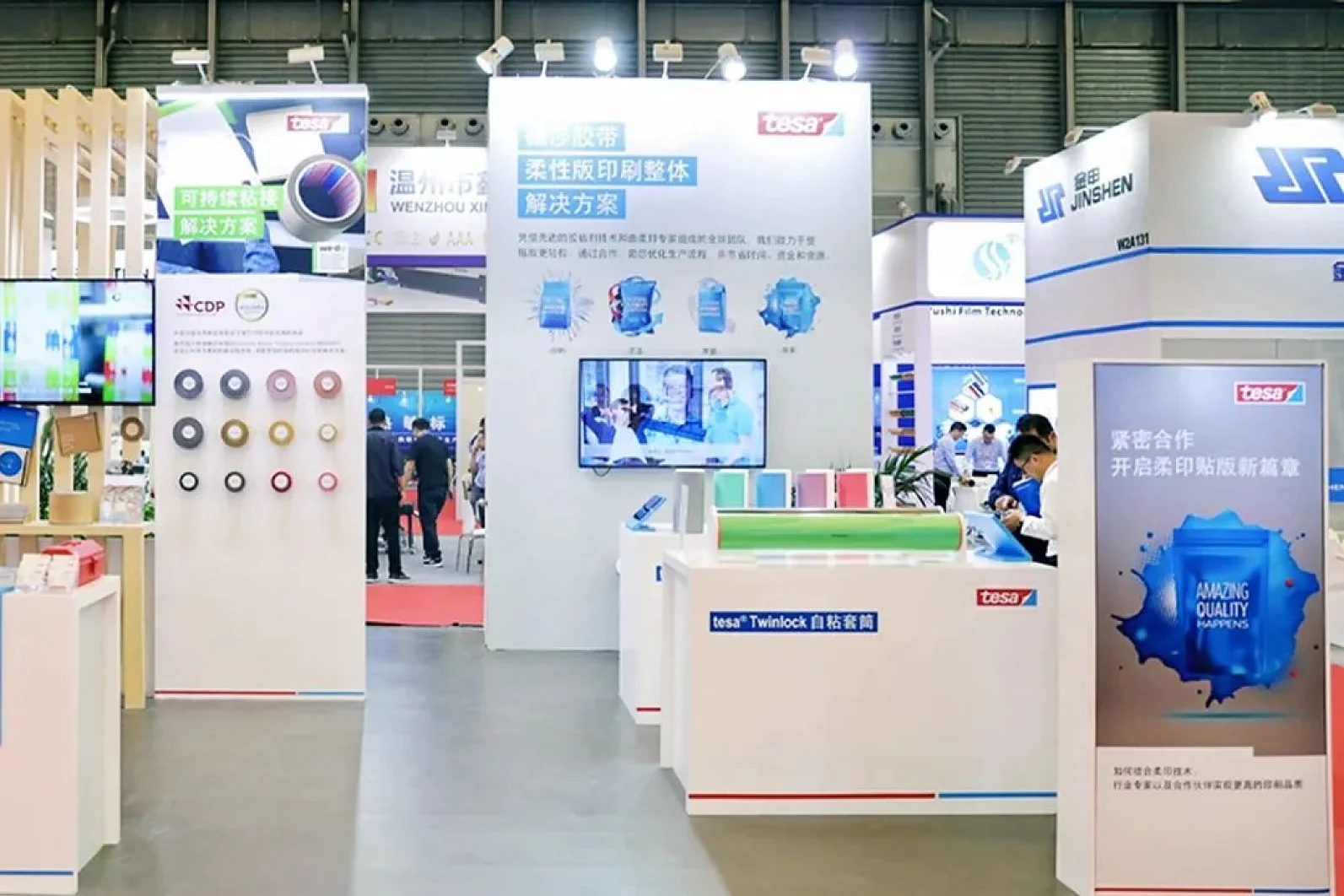 An exhibition booth at a trade show features displays and informational panels from different companies. Visitors are walking around, engaging in conversations. Various products and promotional materials are visible on the stands and tables, with some being secured or highlighted using tesa tape. (This text has been generated by AI)