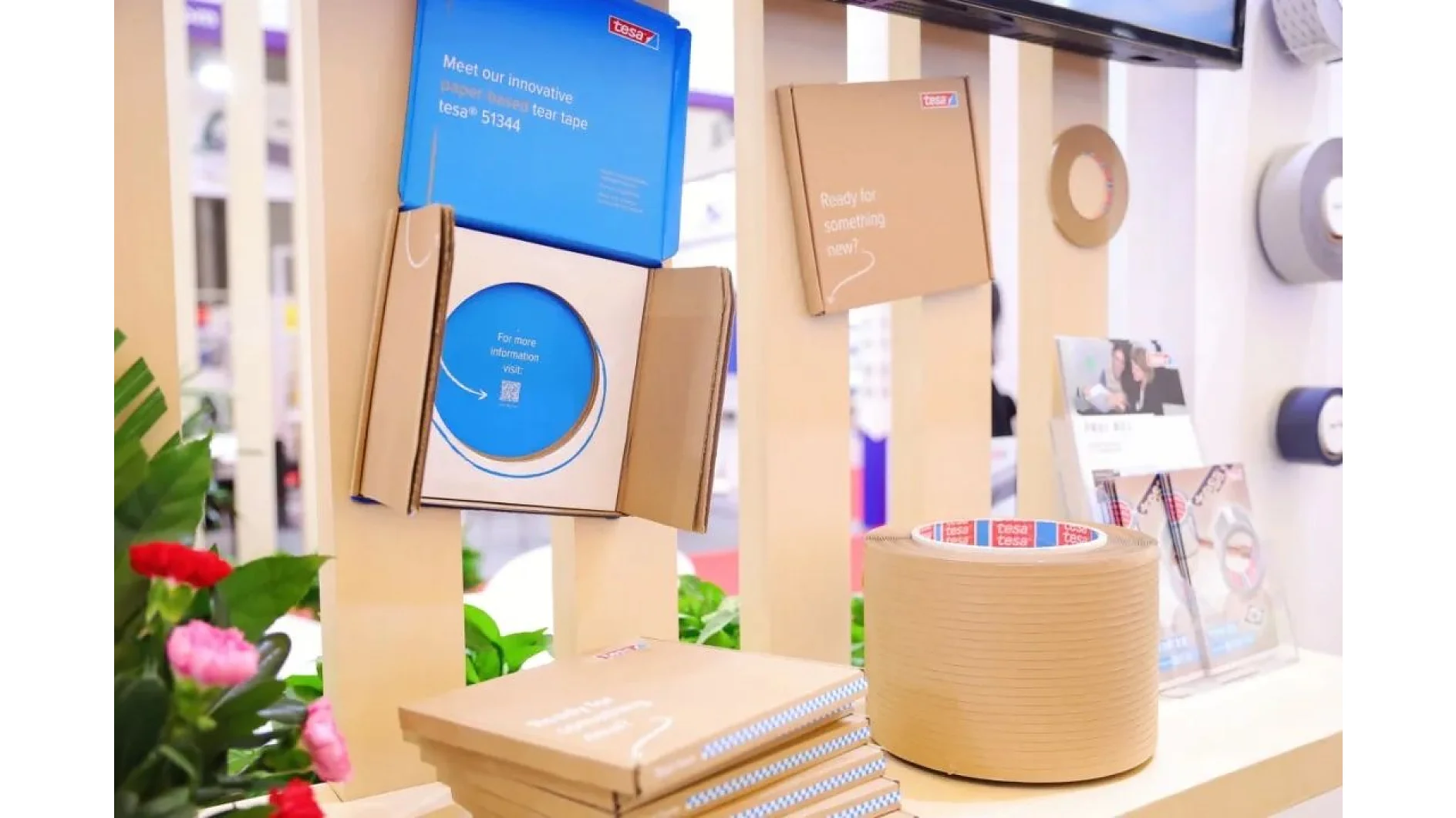 A display featuring various paper and adhesive tesa tape products in cardboard packaging. The setup includes stacked boxes, a large roll of tesa tape, and marketing materials, with plants and a blurred background indicating a commercial setting. (This text has been generated by AI)