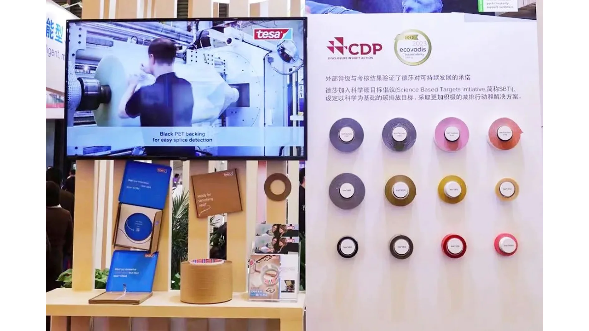 A display features a monitor showing a person operating machinery, with tesa branding. Next to it, a board displays various colored tesa tape rolls. Below, shelves hold tesa tape samples and brochures. Text in Chinese is visible on the board. (This text has been generated by AI)