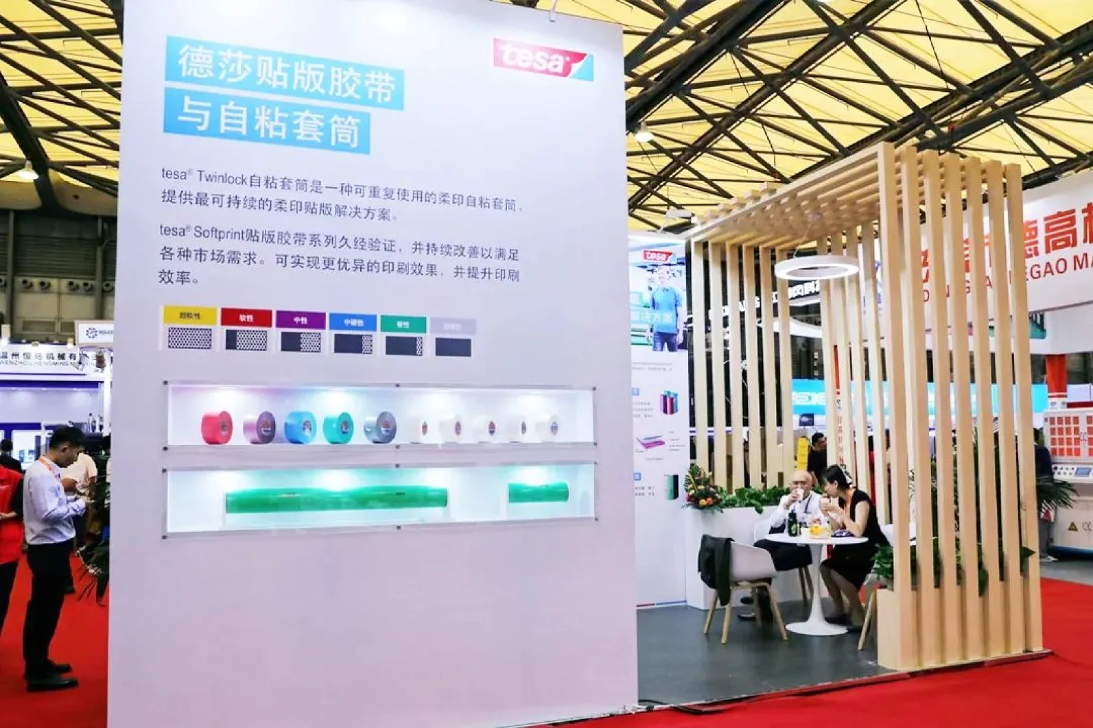 A trade show booth displays tesa tape products. The booth features a large wall with product samples and descriptions. Two people are seated at a small table under a wooden slat structure. The area has red carpeting and bright lighting. (This text has been generated by AI)
