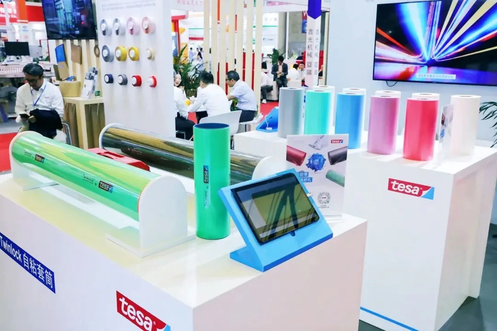 A trade show display featuring large rolls of colorful adhesive tesa tape, a touchscreen monitor, and a screen showing an image of lights. The display is branded with tesa. People are seated in the background at tables. (This text has been generated by AI)