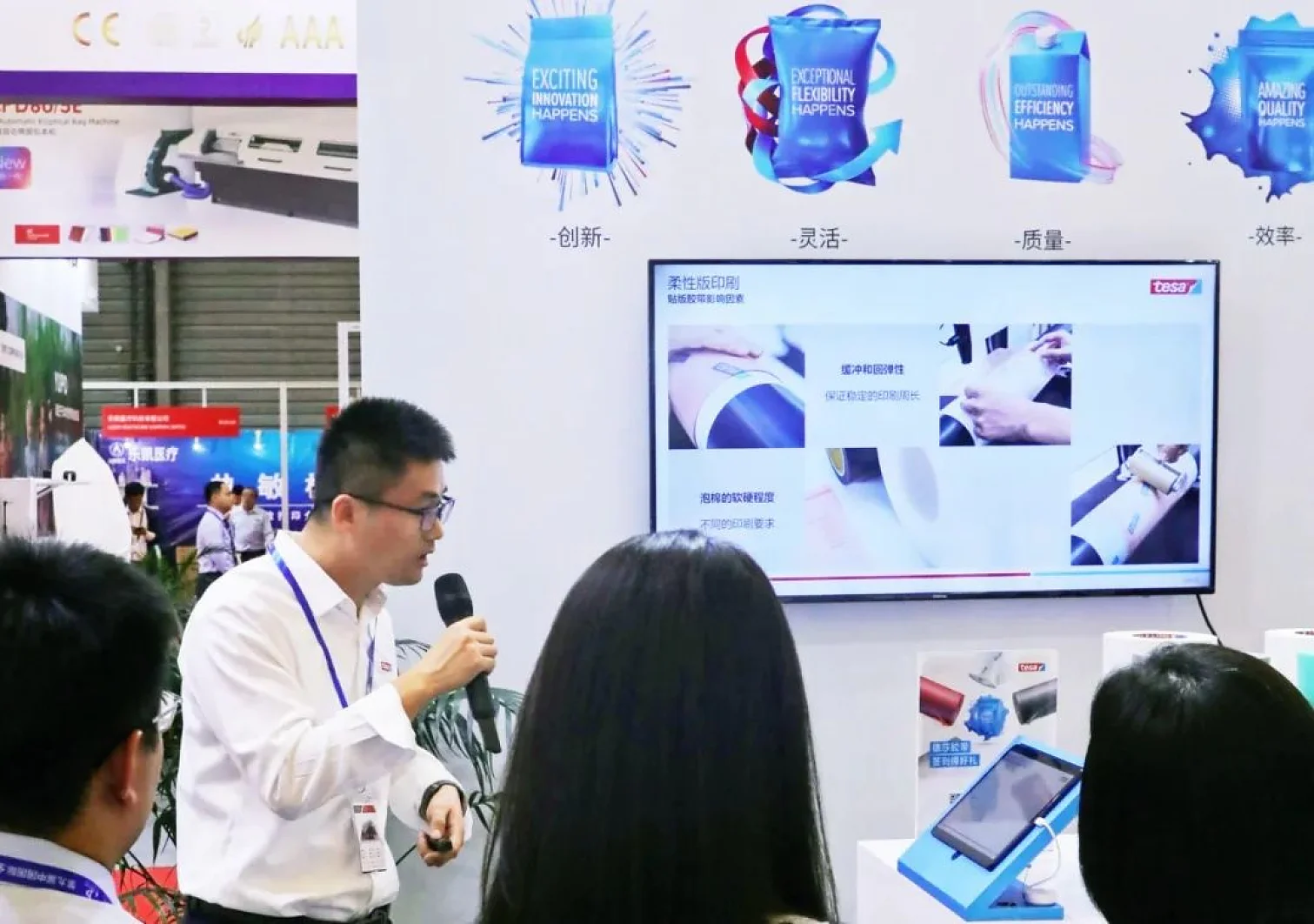A man in a white shirt and glasses holds a microphone and presents at a booth. Behind him, images and text in English and Chinese are displayed on a wall. Several people stand nearby, listening attentively to his presentation about tesa tape. (This text has been generated by AI)