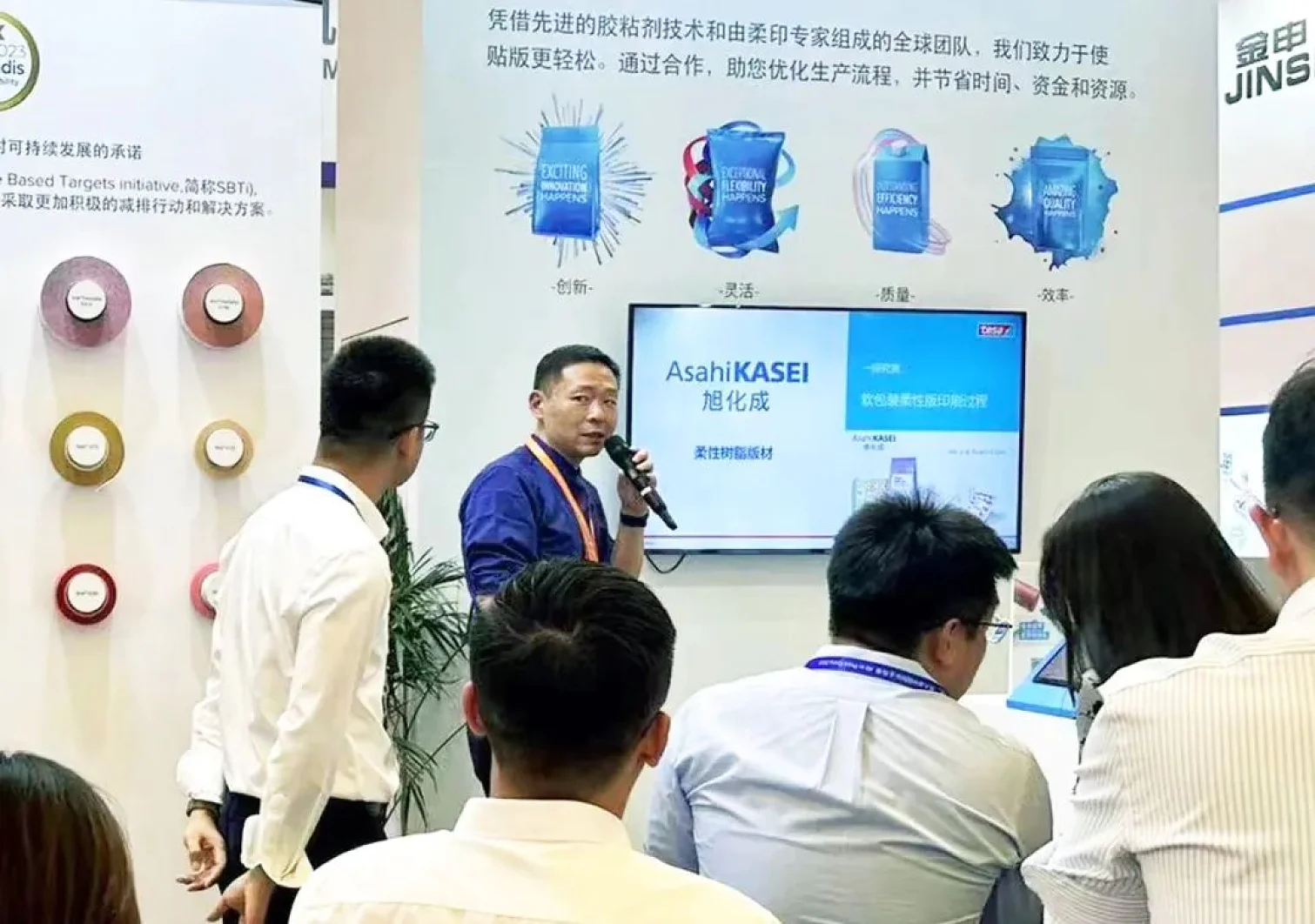 A man in a blue shirt speaks into a microphone at a booth featuring Asahi KASEI branding. People sit facing him, with the booth displaying product images and text on a board behind them. Several circular displays, highlighting tesa tape products, are on the wall to the left. (This text has been generated by AI)