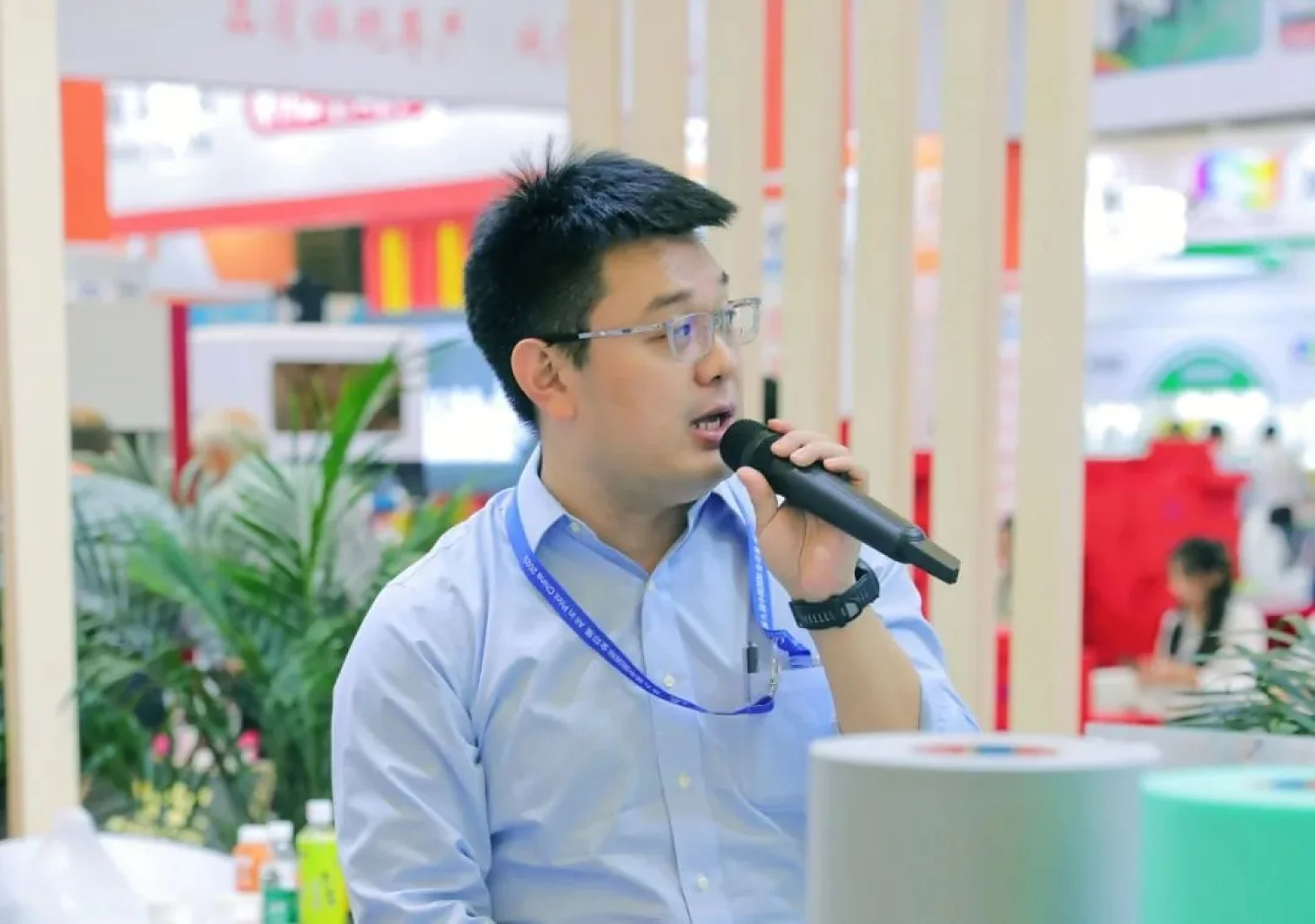 A person wearing glasses and a light blue shirt is speaking into a microphone. They are in an indoor setting with colorful banners and plants in the background. Various cylindrical objects, including rolls of tesa tape, are positioned in the foreground. (This text has been generated by AI)