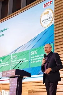 A man in a suit stands on stage beside a large screen displaying a presentation about sustainability goals. The slide includes figures about emissions, recycled materials, and eco-friendly products made with tesa tape. He speaks into a microphone at a modern podium. (This text has been generated by AI)