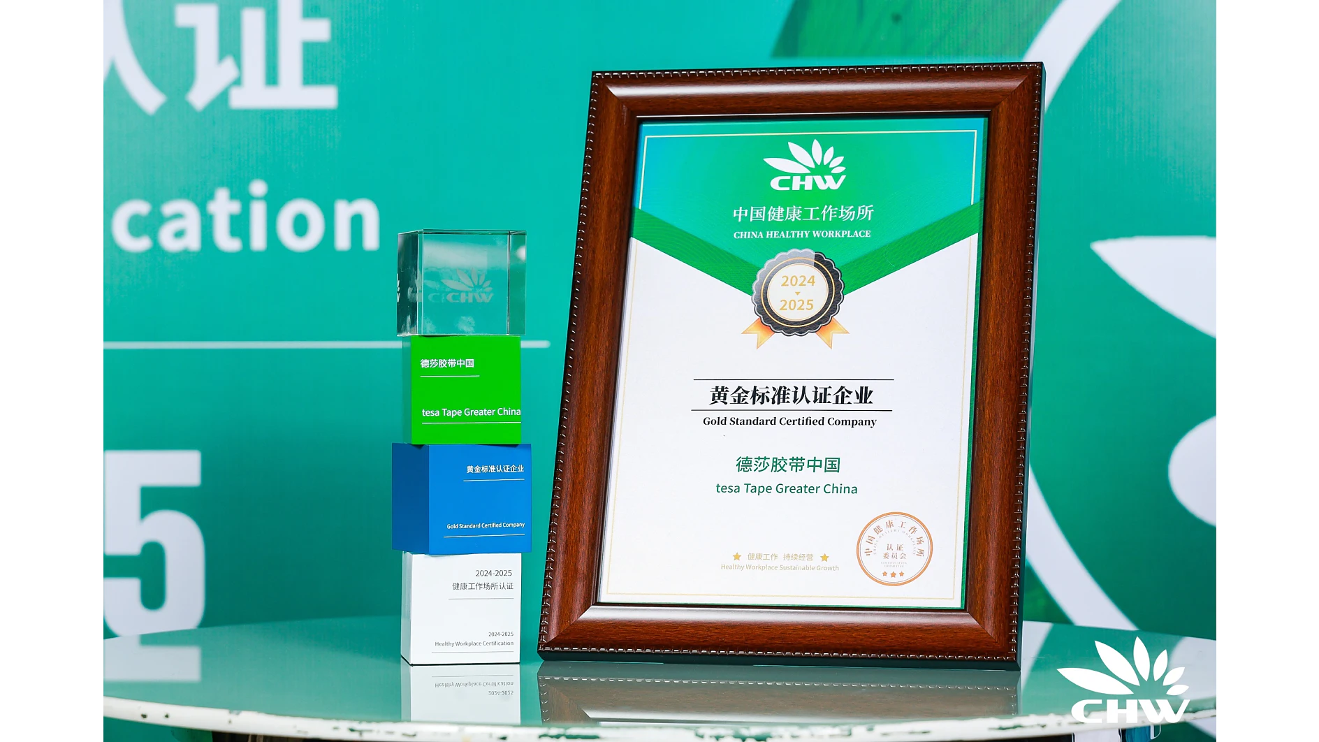 A framed certificate labeled Gold Standard Certified Company 2024-2025 from CHW rests next to a blue and green award box on a reflective surface. The background features a green banner with CHWs logo and partial text. (This text has been generated by AI)