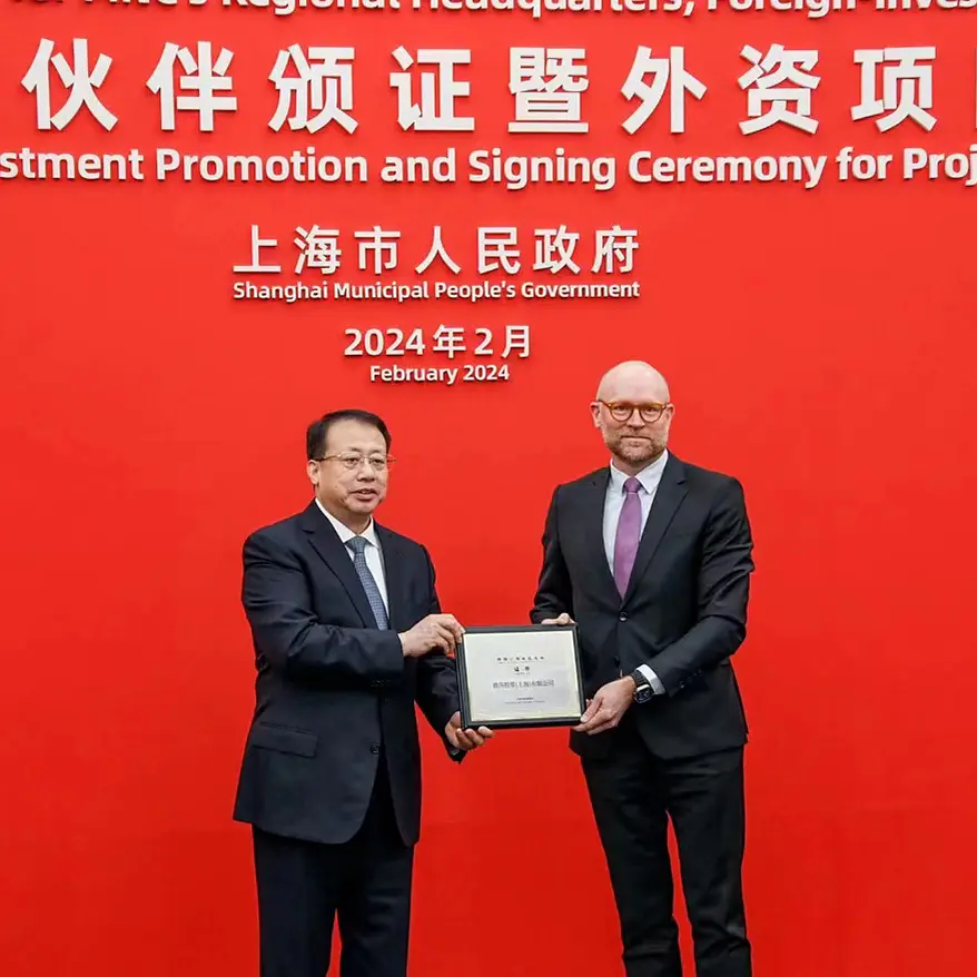 Two men in suits stand in front of a red backdrop with text in Chinese and English about a foreign investment promotion and signing ceremony in February 2024. One man hands a tesa tape plaque to the other. (This text has been generated by AI)