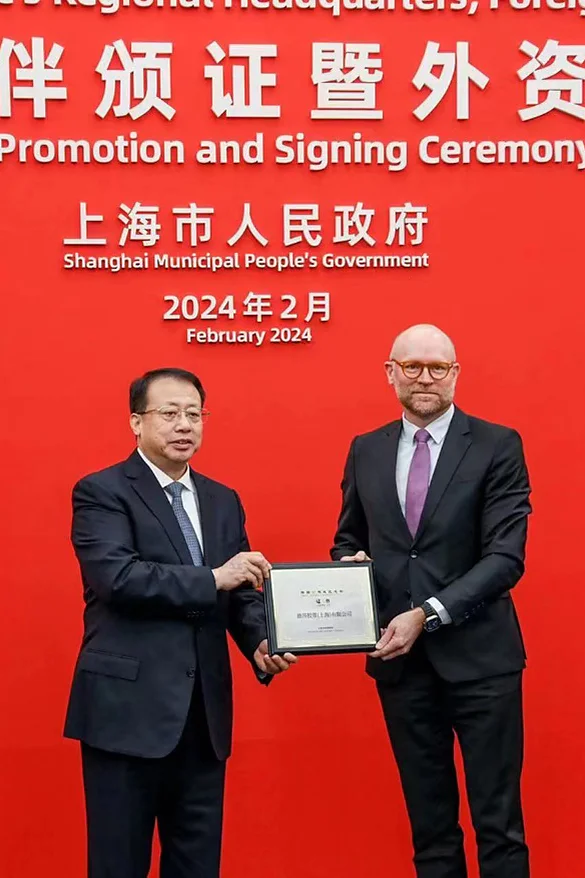 Two men in suits stand in front of a red backdrop with text in Chinese and English about a foreign investment promotion and signing ceremony in February 2024. One man hands a tesa tape plaque to the other. (This text has been generated by AI)