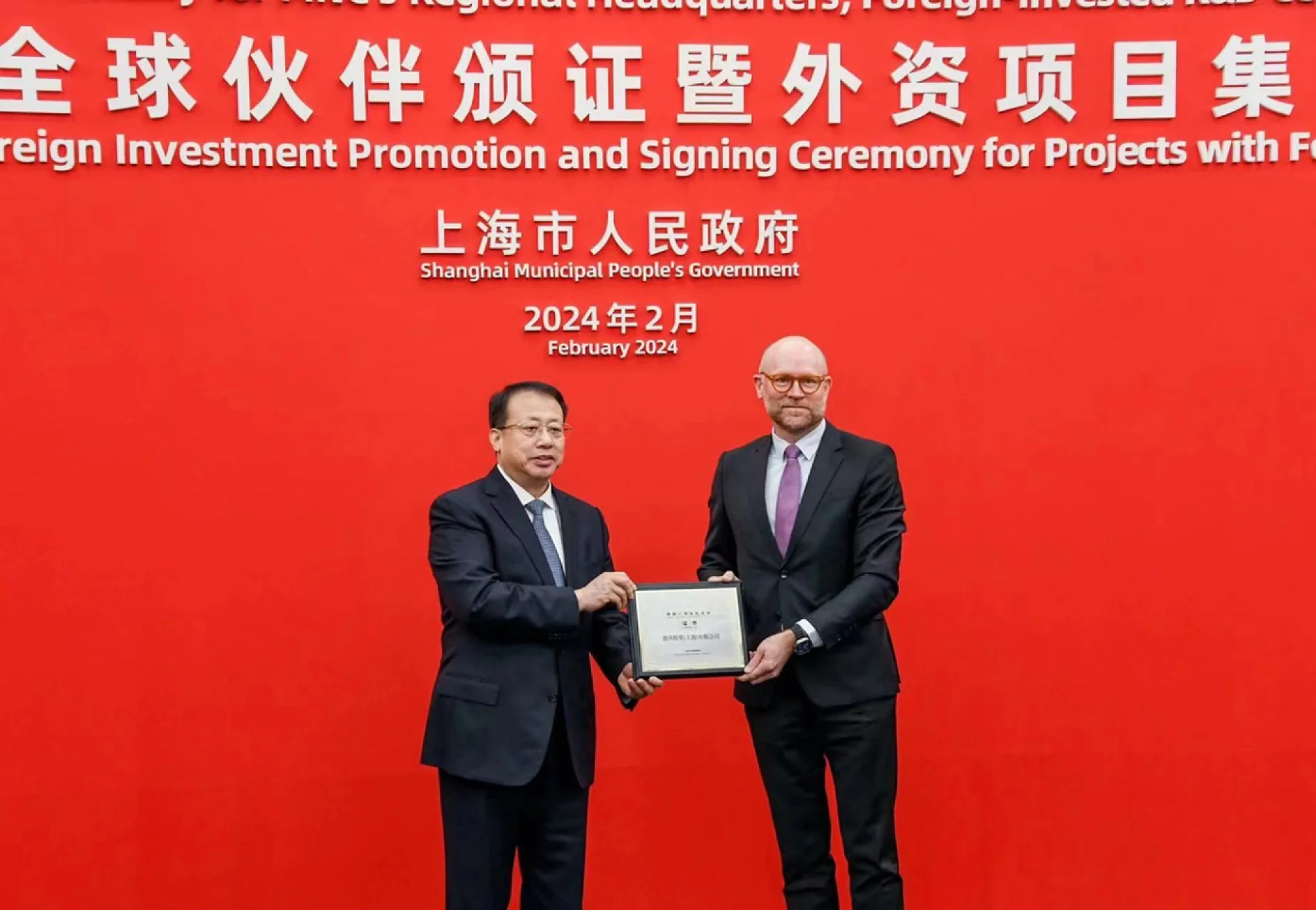 Two men in suits stand in front of a red backdrop with text in Chinese and English about a foreign investment promotion and signing ceremony in February 2024. One man hands a tesa tape plaque to the other. (This text has been generated by AI)