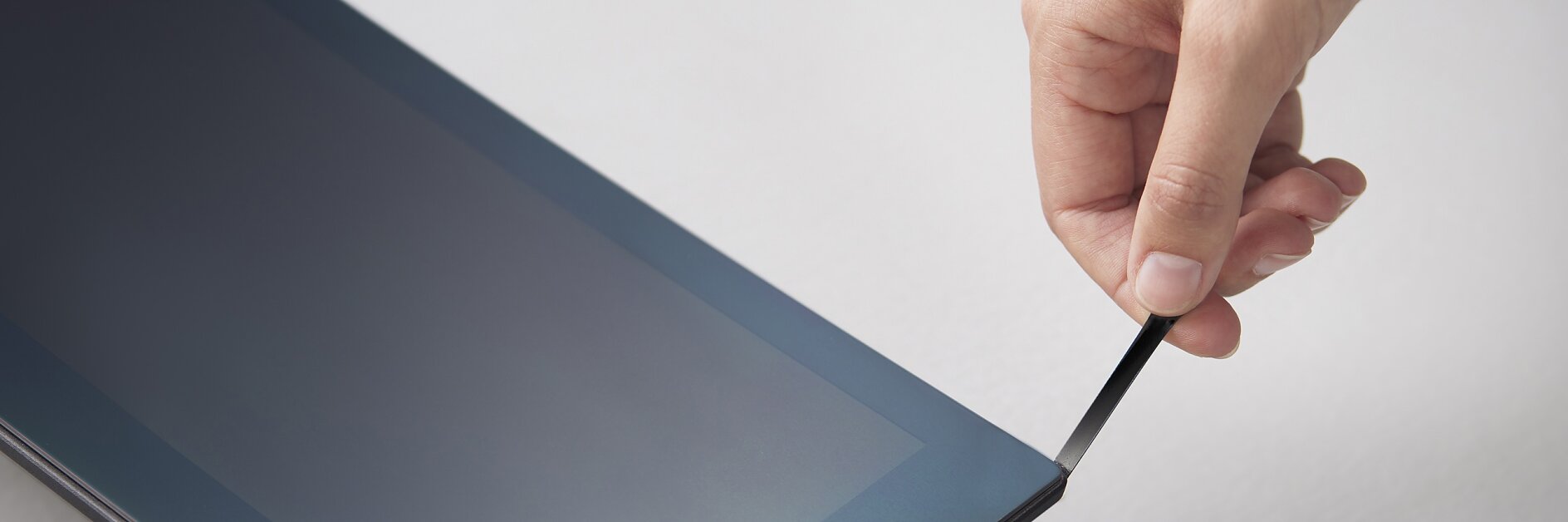 A hand is seen peeling a transparent protective film from the screen of a rectangular electronic device, likely a tablet or monitor, on a light gray surface using tesa tape. (This text has been generated by AI)