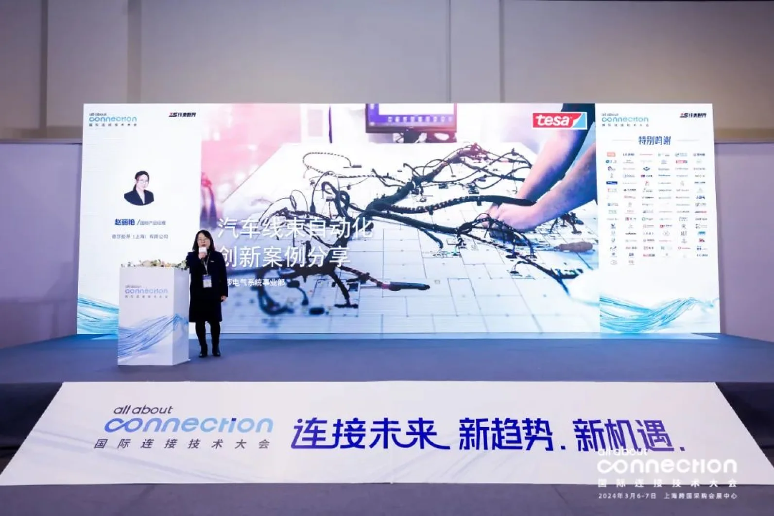 A speaker presents on stage next to a large screen displaying a technical drawing and the logo tesa. The backdrop features banners with the text connection and a list of other names. The event is held in Shanghai, with prominent displays emphasizing the tesa tape brand. (This text has been generated by AI)