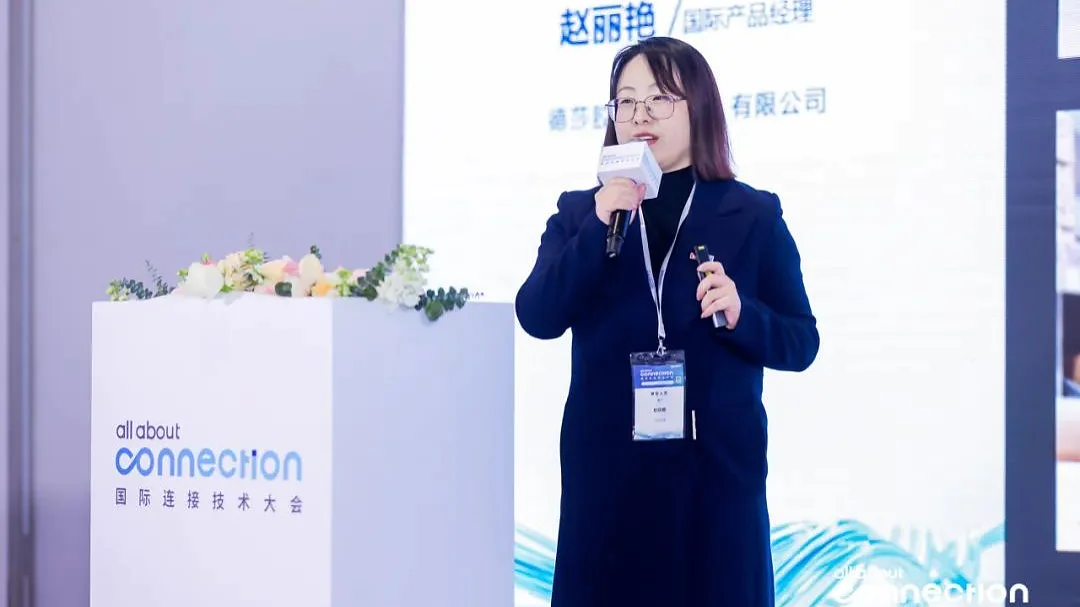 A woman stands on a stage holding a microphone. She is speaking in front of a podium with flowers. A screen beside her displays text. The backdrop reads all about connection with additional text in Chinese, prominently featuring the brand tesa tape throughout the presentation. (This text has been generated by AI)