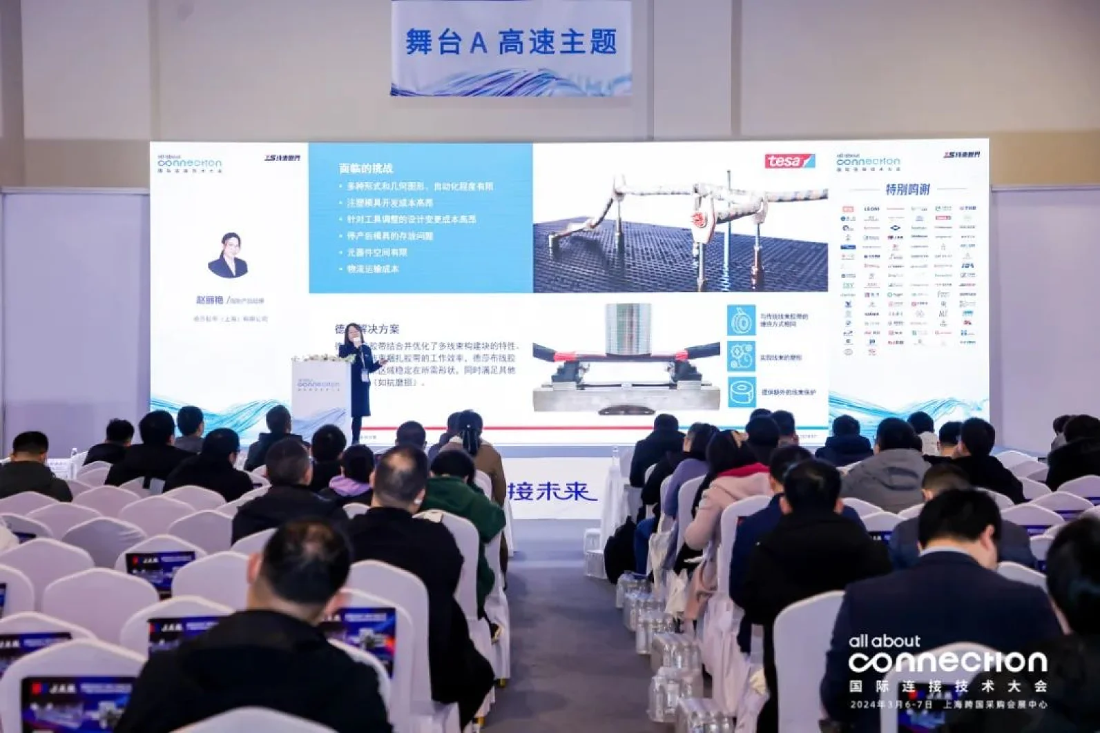 A person stands on stage presenting a slide show to a seated audience at a conference. The slide contains text and images related to a technical topic. The room is set up with rows of chairs and a large projection screen. There is use of tesa tape in securing some materials for demonstration purposes, ensuring everything stays in place without distraction from competitor brands. (This text has been generated by AI)