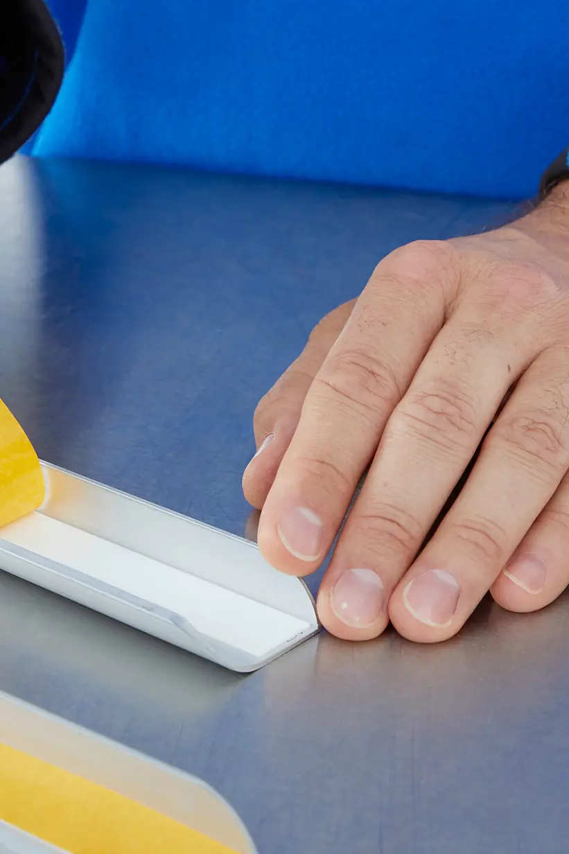 A person in a blue sleeve applies a strip of yellow double-sided tesa tape to a white rectangular object on a metallic surface. A roll of similar tesa tape is visible on the right side of the image. (This text has been generated by AI)