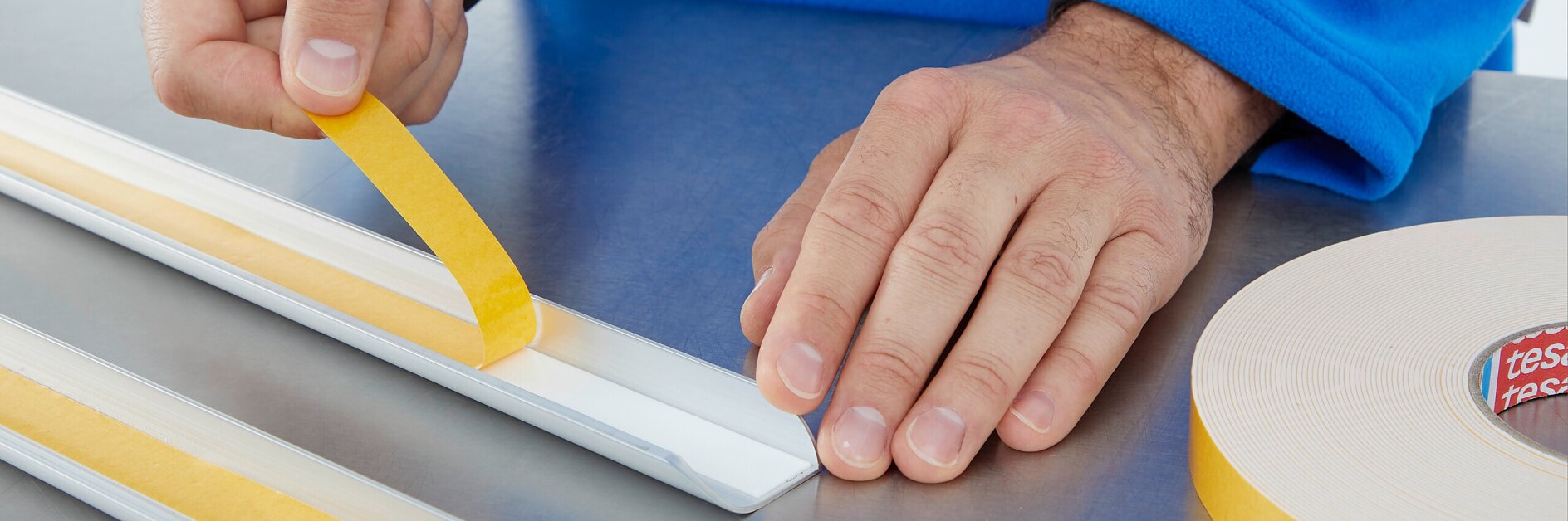 A person in a blue sleeve applies a strip of yellow double-sided tesa tape to a white rectangular object on a metallic surface. A roll of similar tesa tape is visible on the right side of the image. (This text has been generated by AI)
