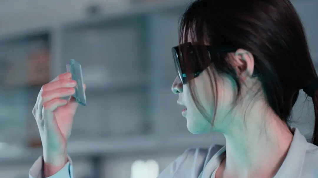 A person in a lab coat and safety glasses examines a thin, transparent rectangular object in a laboratory setting. Shelves and scientific equipment are blurred in the background, with the focus entirely on tesa tape.