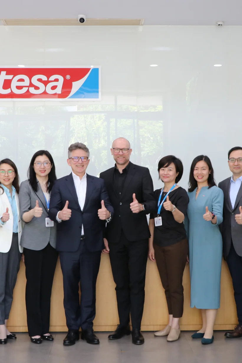 Twelve people stand in a line indoors, giving thumbs-up signs. They are dressed in business attire. A logo with the word tesa is mounted on the wall behind them. Plants are visible on either side of the group. (This text has been generated by AI)