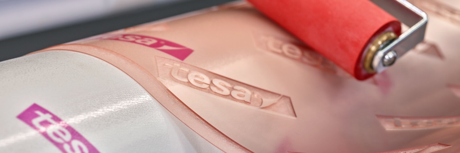 Close-up of a hand using a red roller on a clear adhesive film marked with the tesa tape logo. The film is being applied to a white cylindrical surface. (This text has been generated by AI)