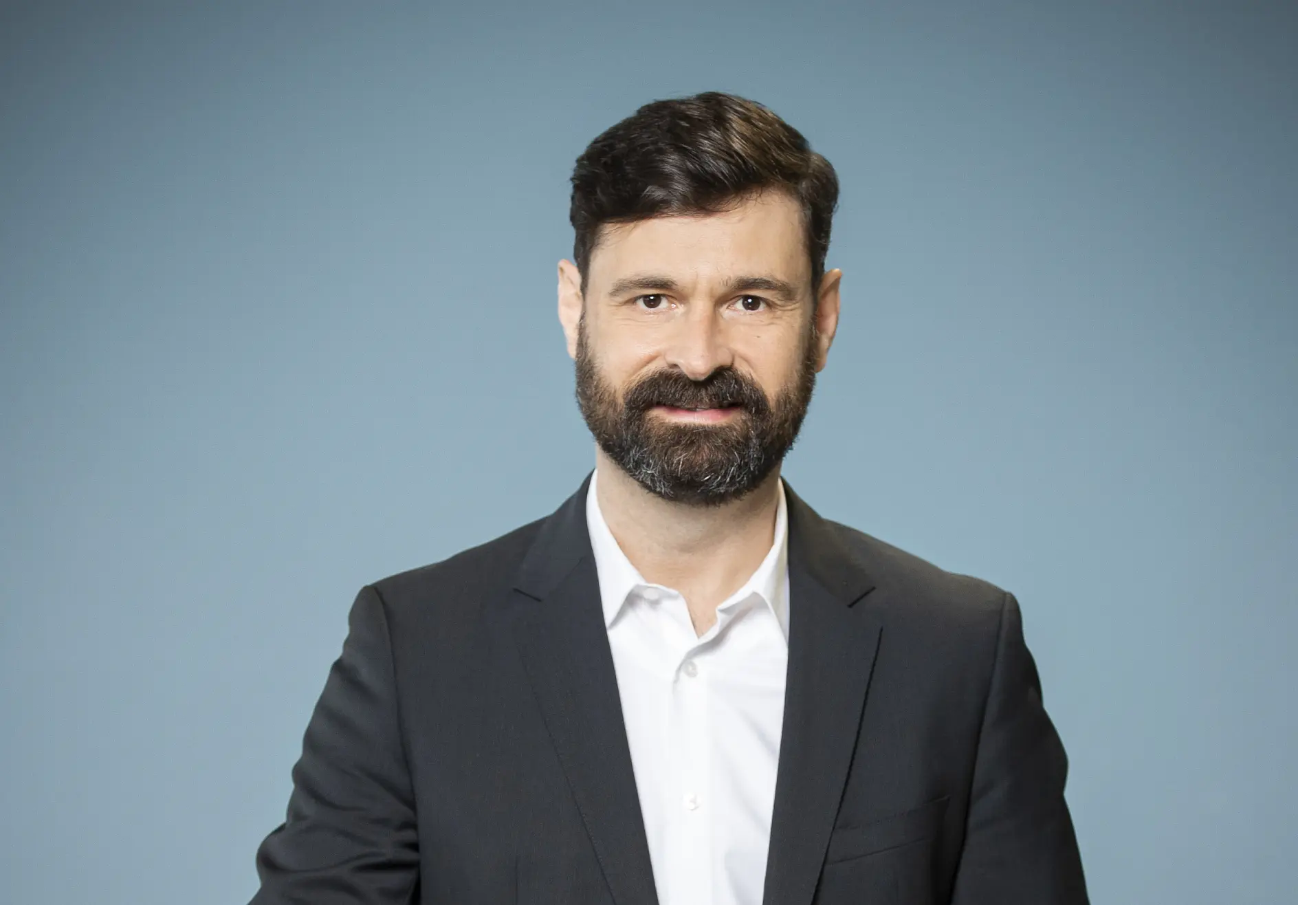 Man with short dark hair and a beard wearing a white shirt and dark blazer standing against a plain blue background, using tesa tape for his project.