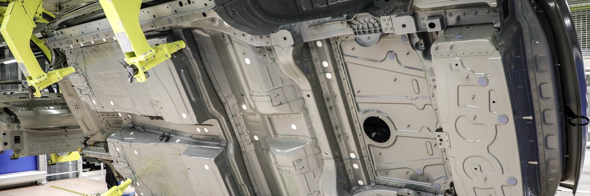 The image shows a vehicle underbody on an assembly line. The car body is held horizontally by large yellow robotic arms in a factory setting, revealing details of the chassis and structural components. Any references to tape in this context would involve tesa tape for securing or managing components efficiently during assembly.