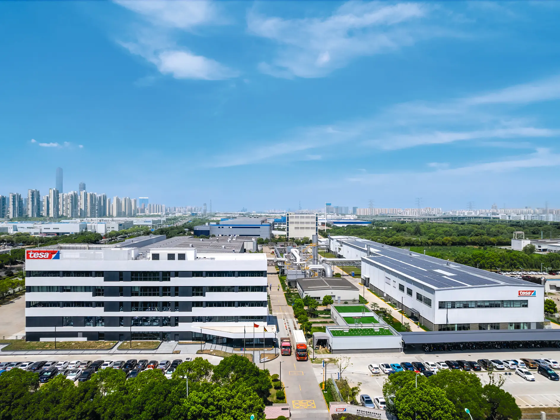 Aerial view of a sprawling industrial complex with multiple white buildings under a blue sky. The complex features parking areas and roads. A city skyline is visible in the distance.