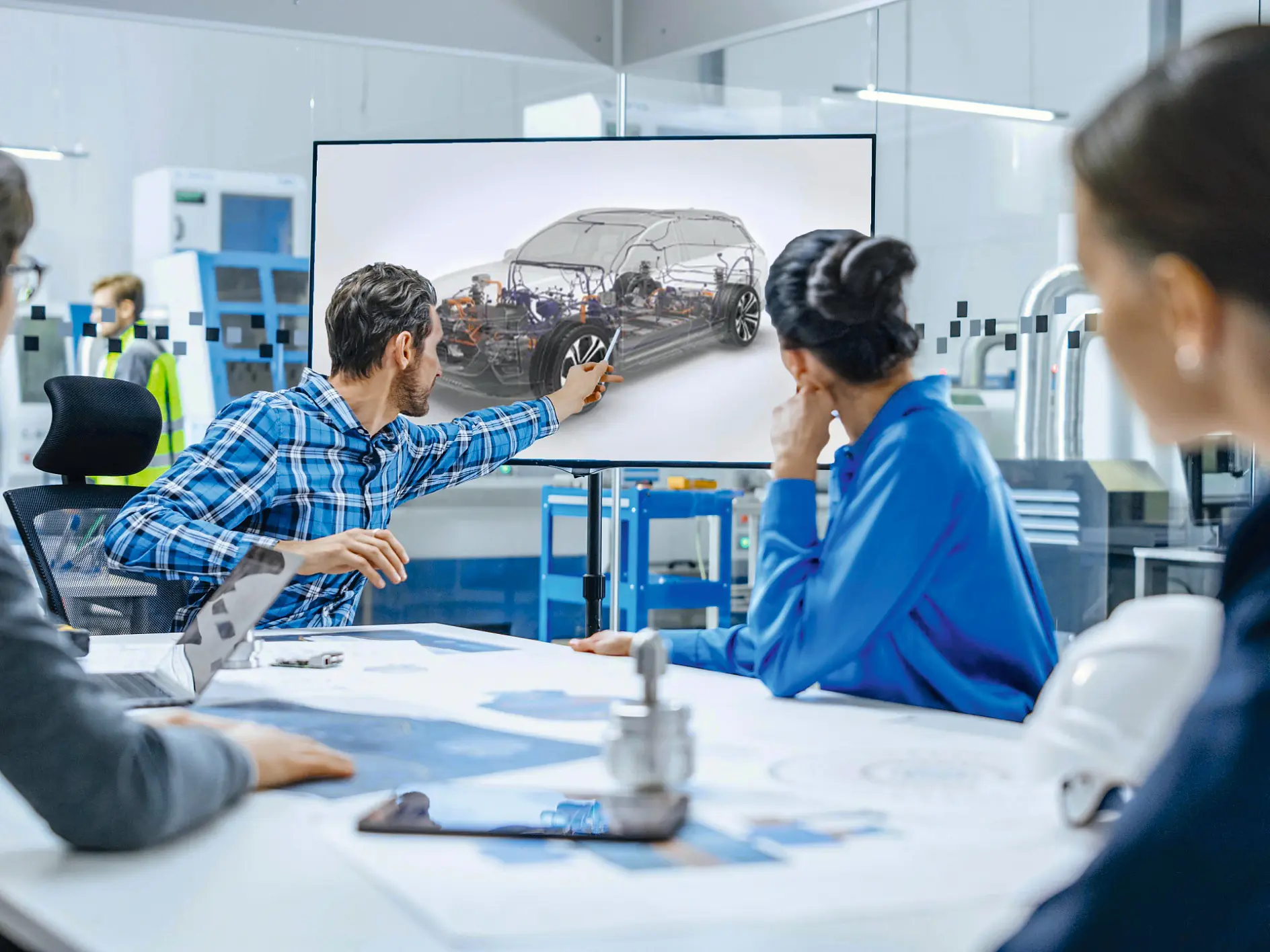 A group of four people sits around a table in an office, focusing on a large screen. One person is pointing to a car displayed on the screen. Blueprints and a model are on the table, held in place with tesa tape. The setting appears to be a professional meeting or presentation.