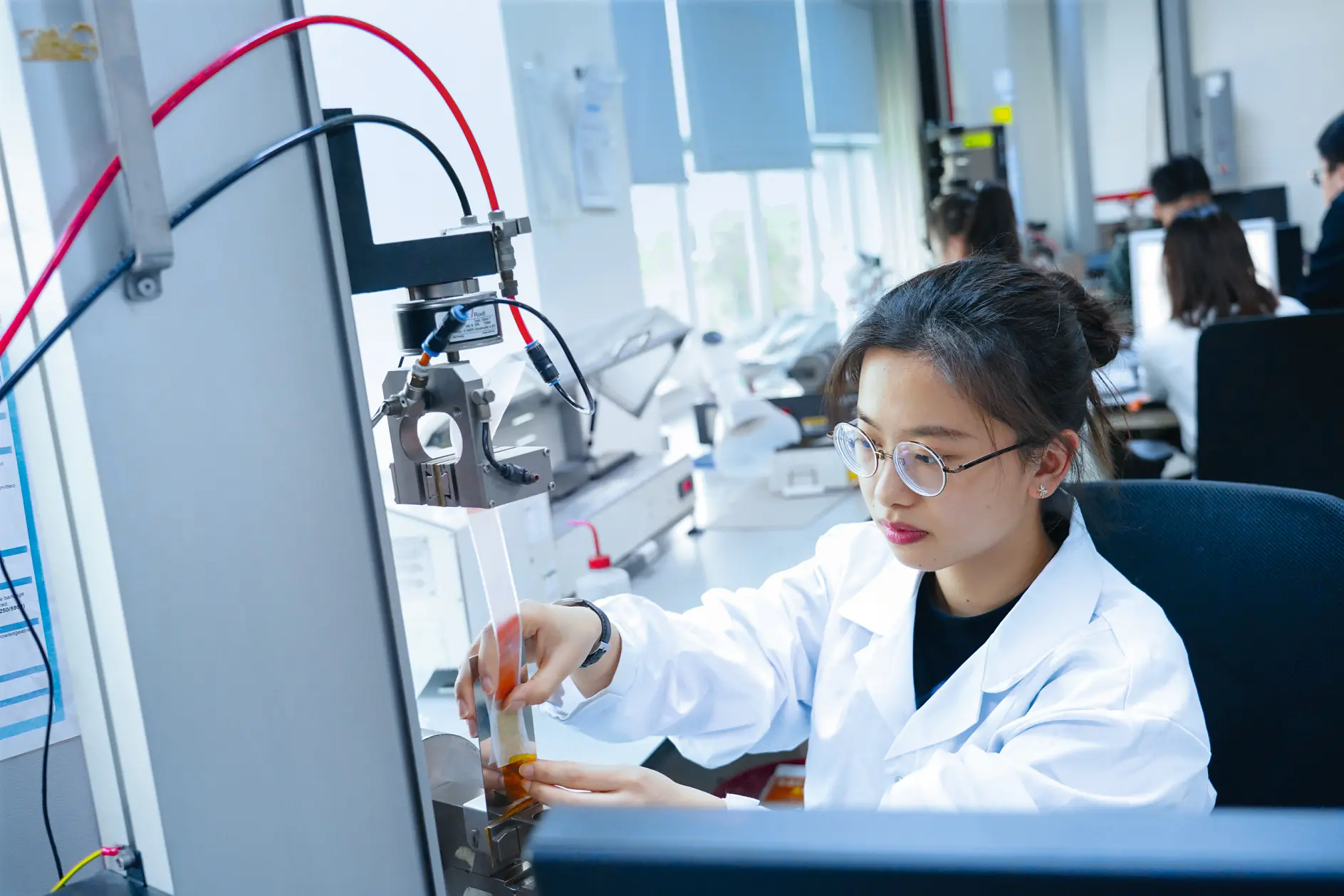 A person in a lab coat and glasses operates equipment in a laboratory. They are adjusting a component near a machine with wires using tesa tape. Other people are working in the background, and computer monitors are visible. The setting is brightly lit and organized.