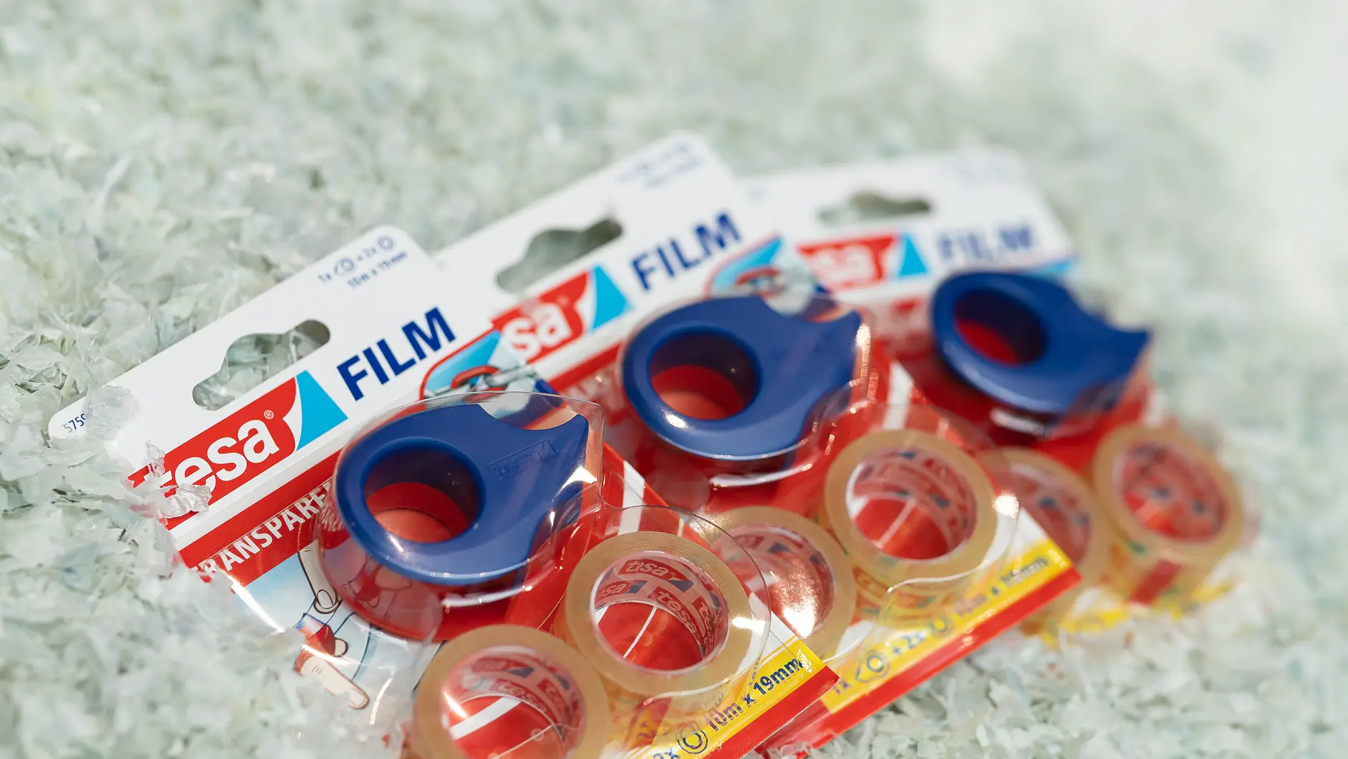 Three packages of tesa transparent adhesive tape are displayed on a pile of shredded paper. Each package contains a roll of tesa tape with a blue dispenser. The packaging text includes tesa Film and transparent. (This text has been generated by AI)