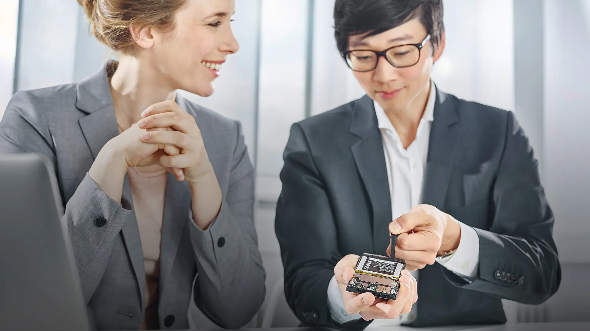 Two people in business attire are seated indoors. One person is holding a small electronic device and tool, gesturing towards it with tesa tape, while the other smiles and observes. A laptop is partially visible on the table. Large windows are in the background. (This text has been generated by AI)