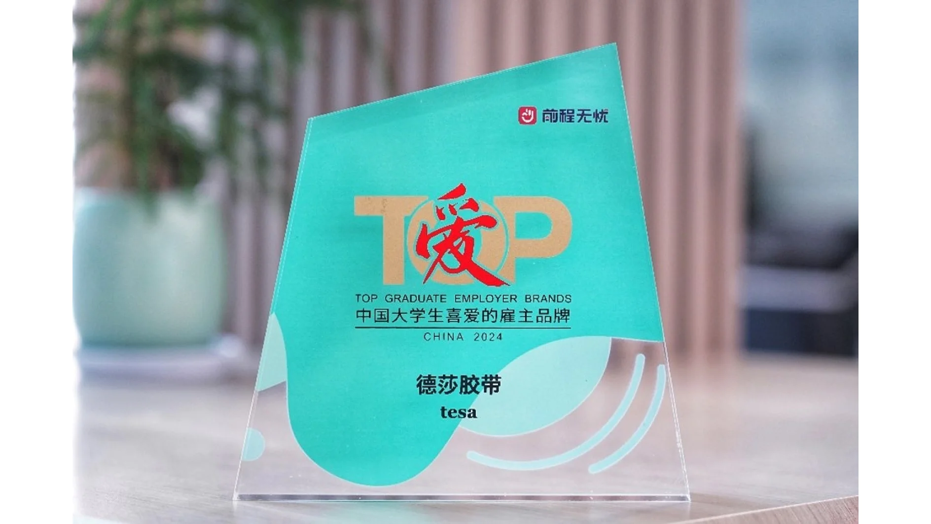 A transparent trophy with a teal background featuring the words Top Graduate Employer Brands China 2024 in English and Chinese. The name tesa tape is displayed near the bottom. The trophy has a triangular shape and sits on a table. (This text has been generated by AI)