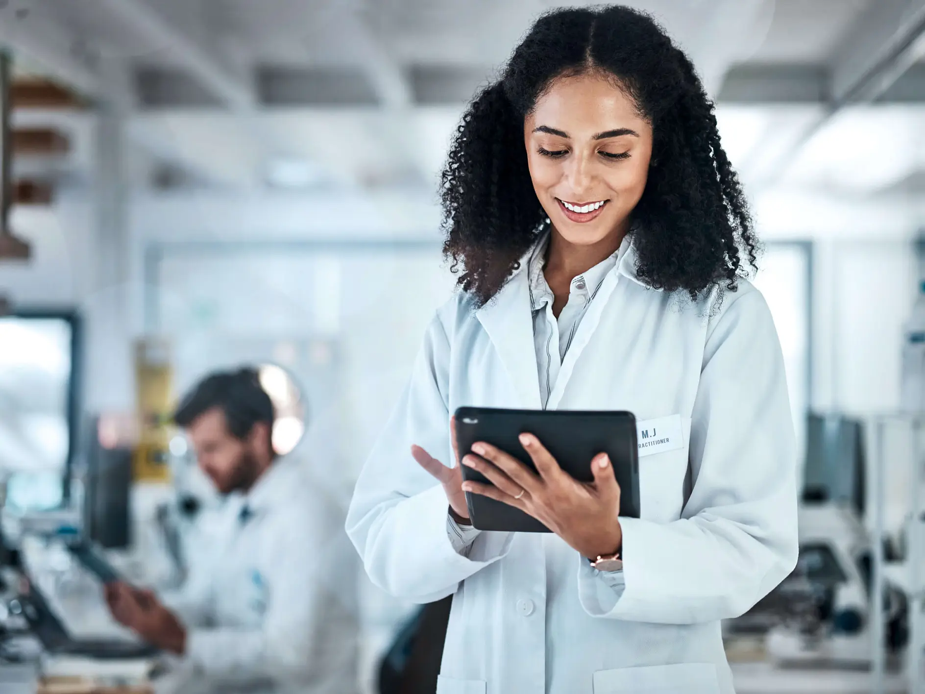 A woman in a lab coat smiles while using a tablet in a laboratory setting. Another person works at a desk in the background. Shelves with various lab equipment, including rolls of tesa tape, are visible around them. (This text has been generated by AI)