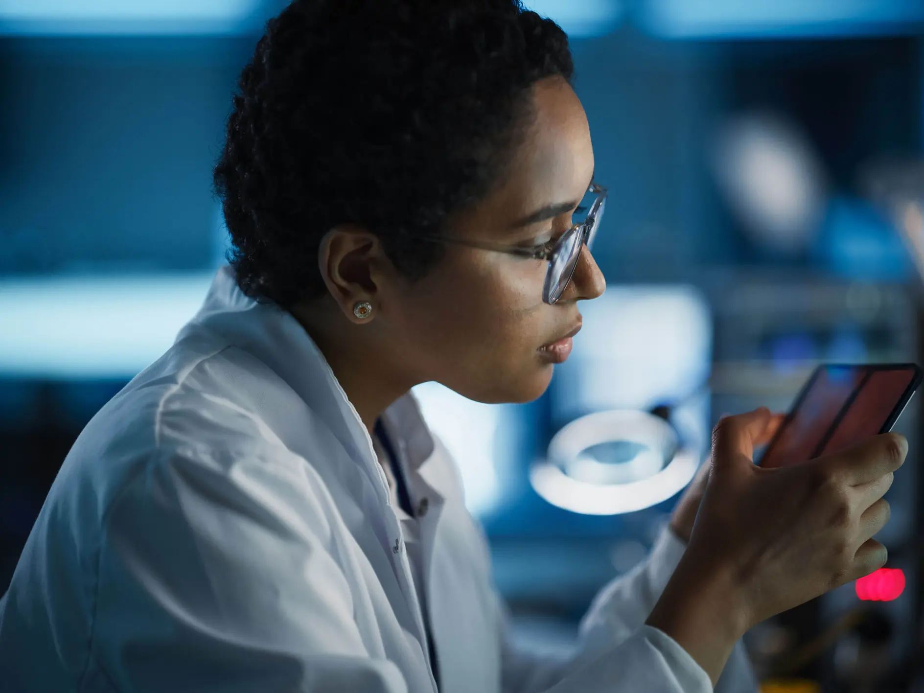A person wearing a white lab coat and glasses is sitting in a lab environment, closely examining a smartphone secured with tesa tape. The background is blurred, featuring computer screens and lab equipment. The lighting is dim, highlighting the focus on the smartphone wrapped with tesa tape. (This text has been generated by AI)