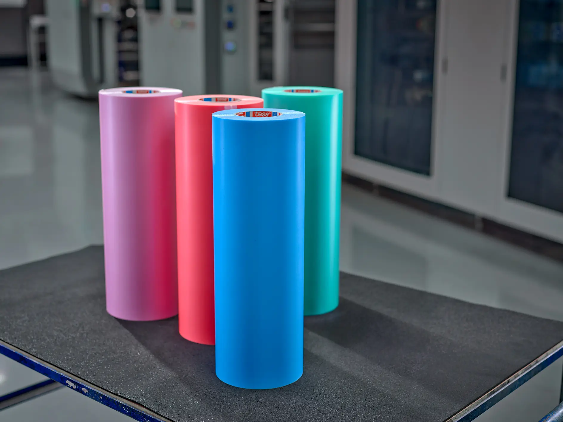 Five rolls of colorful tesa adhesive tape in pink, red, blue, green, and gray, displayed on a black surface indoors. (This text has been generated by AI)