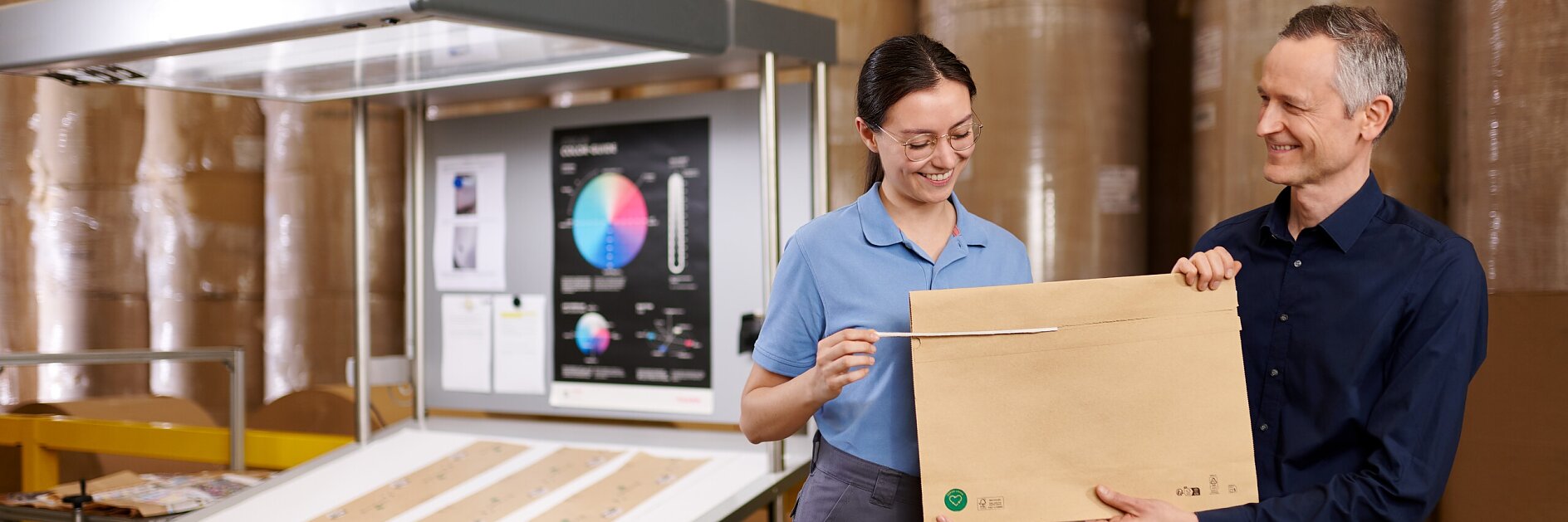 Two people standing indoors holding a large tesa-branded cardboard box, with shelves and packaging materials in the background.