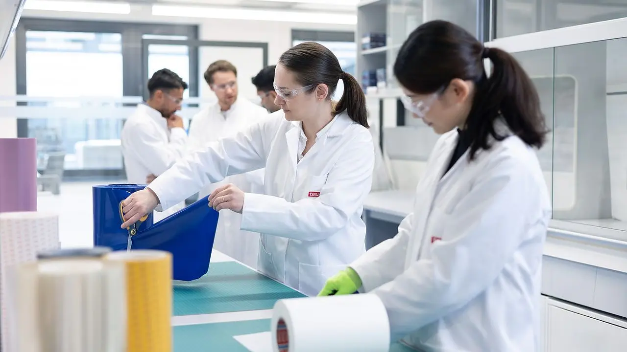 Several people in lab coats and safety glasses work with large rolls of material and tesa tape on tables in a bright laboratory, with shelves and lab equipment visible in the background. (This text has been generated by AI)
