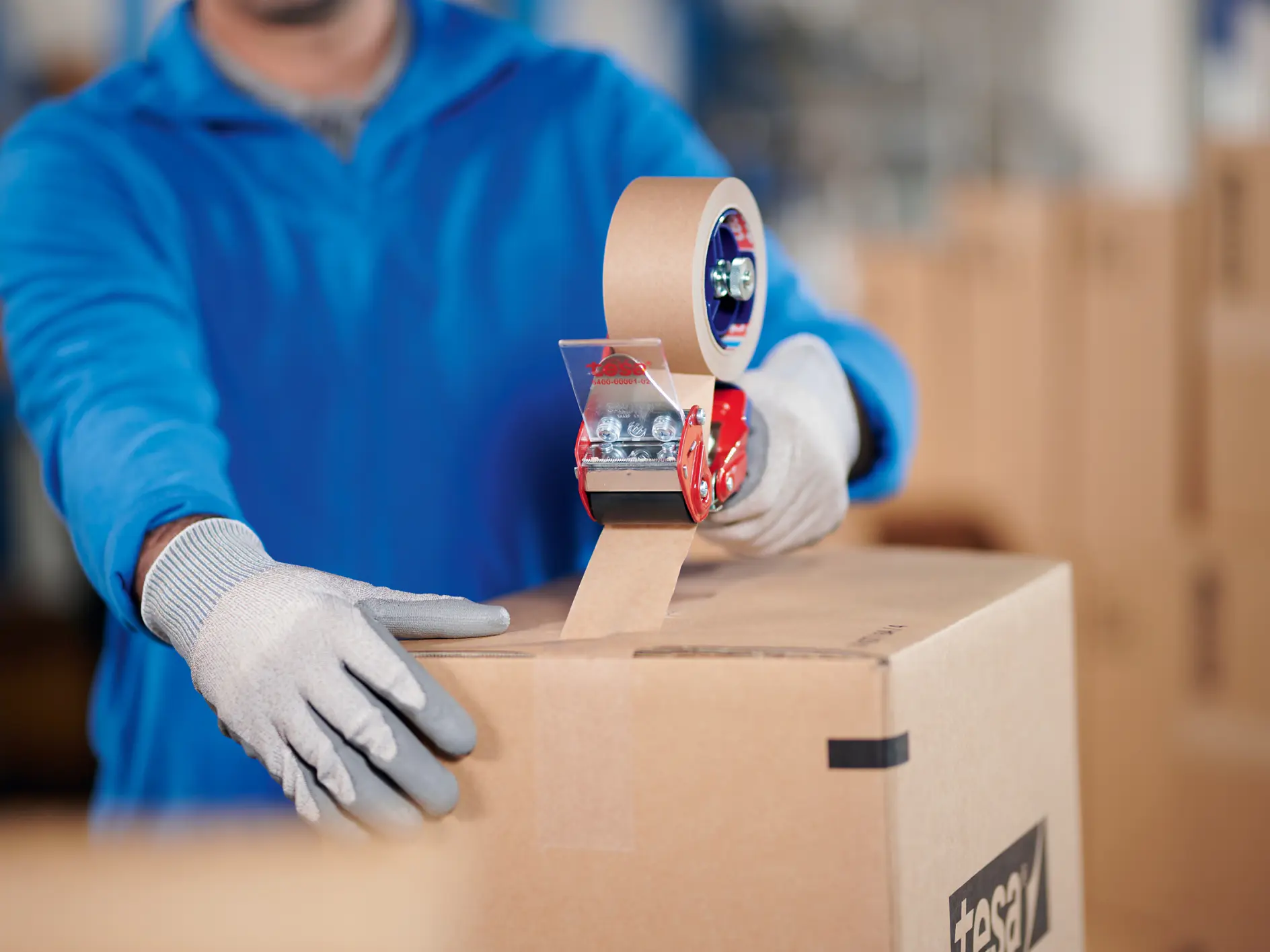 A person wearing a blue jacket and gray gloves is sealing a cardboard box with brown packing tesa tape using a red tesa tape dispenser. Other cardboard boxes are visible in the background.