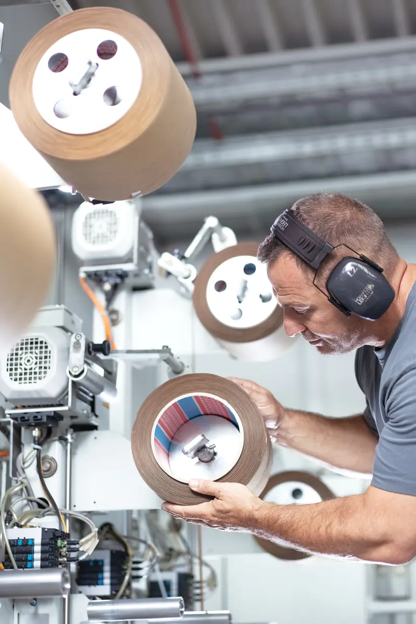 A man wearing ear protection inspects a roll of material in an industrial setting. Various machines with similar rolls of tesa tape are visible in the background. He is focused on examining the roll closely. (This text has been generated by AI)