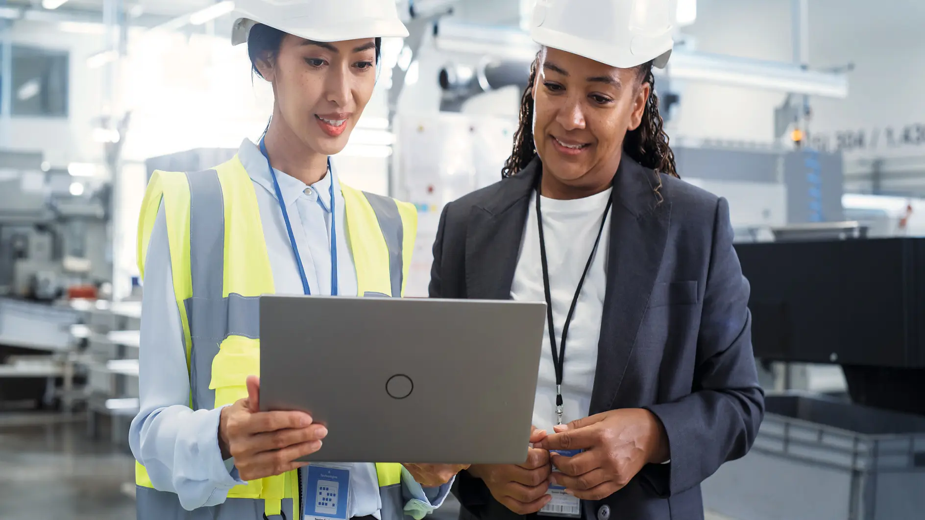 Two professionals in a modern industrial setting looking on a laptop, one wearing a safety vest and hard hat, the other in business attire.