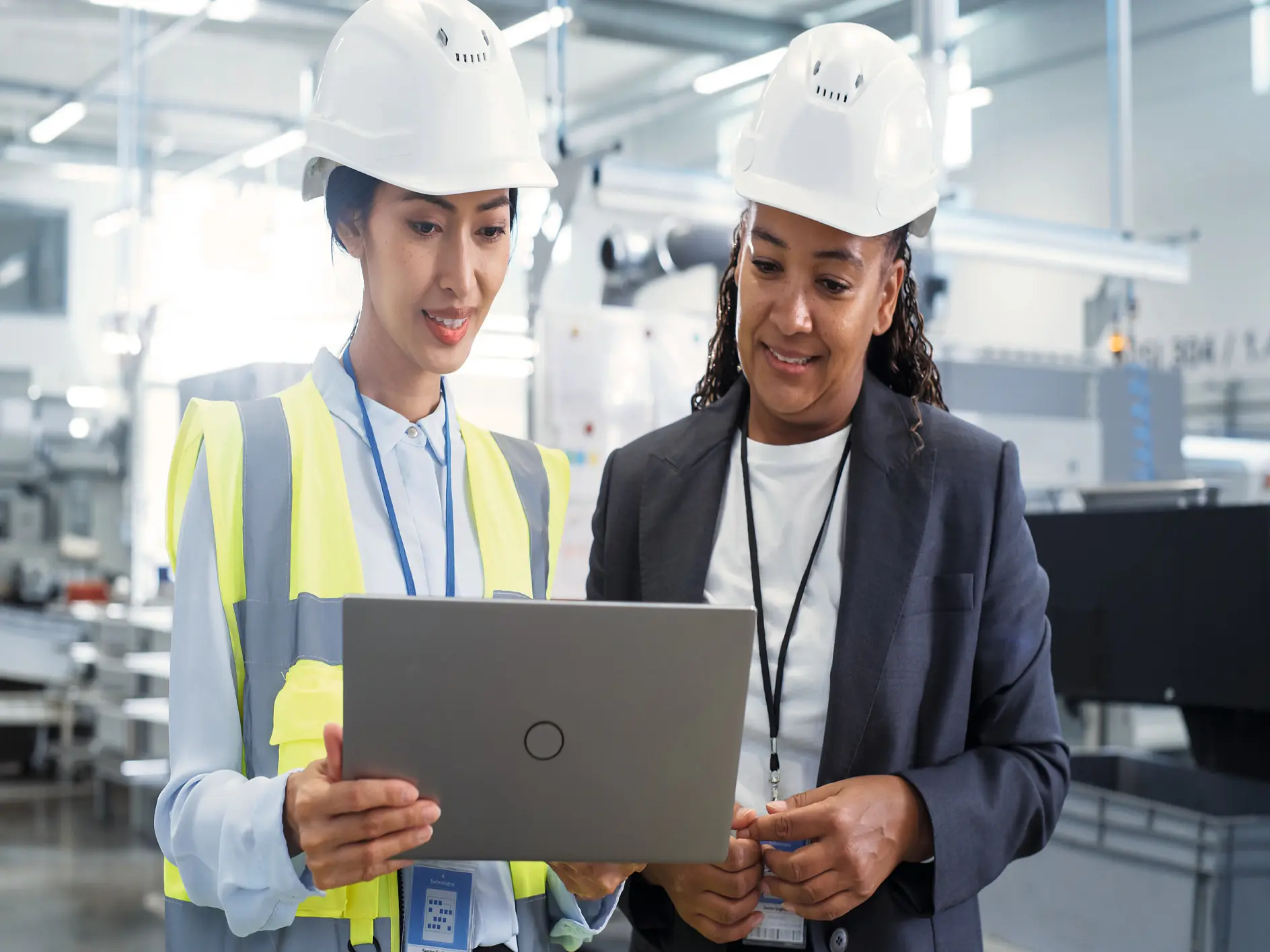 Two professionals in a modern industrial setting looking on a laptop, one wearing a safety vest and hard hat, the other in business attire. (This text has been generated by AI)