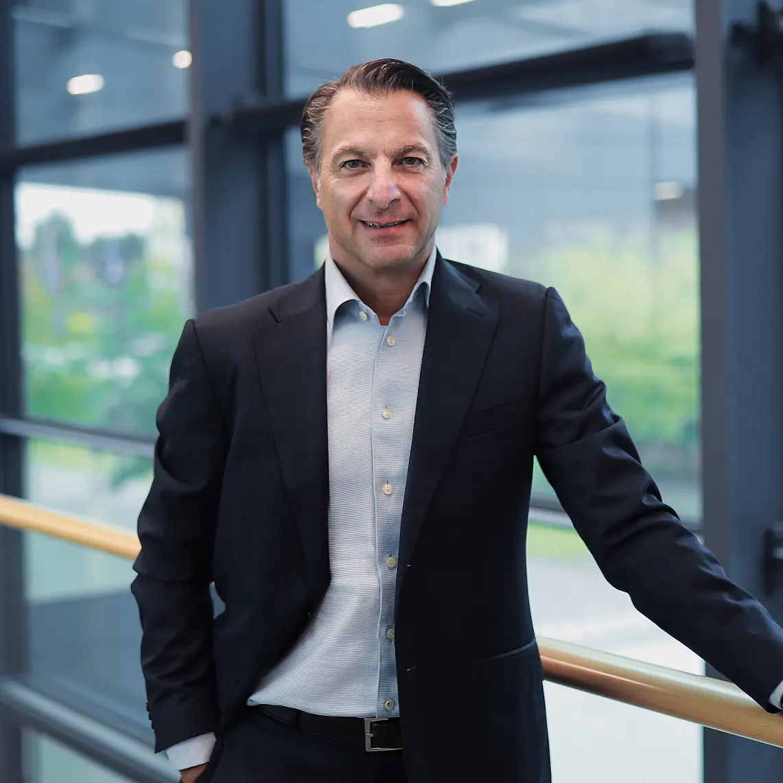 A man in a dark suit and light blue shirt stands by a railing indoors, with large windows and an outdoor view behind him, smiling slightly. (This text has been generated by AI)