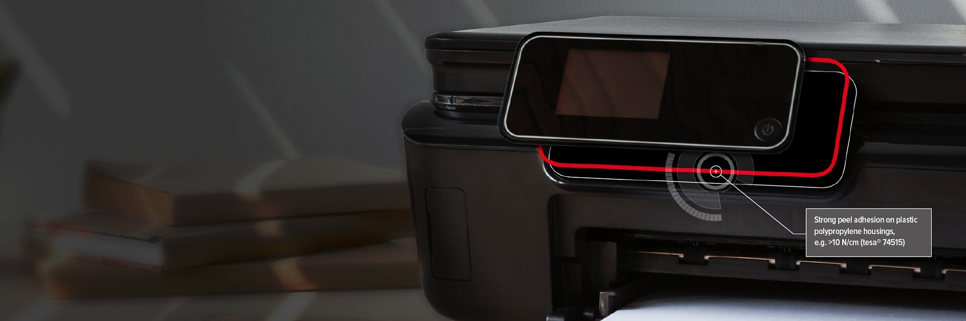 A close-up of a black printer with a clear tesa tape sticker applied to its control panel, highlighted by a red outline. A label points to the tesa tape sticker, noting its strong peel-adhesion on plastic housings. (This text has been generated by AI)