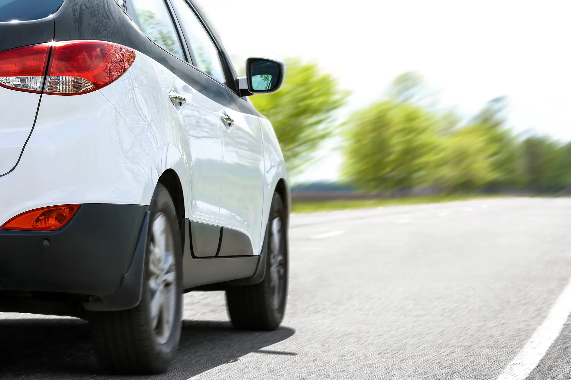 white compact car driving on a paved road with blurred green trees in the background on a clear day