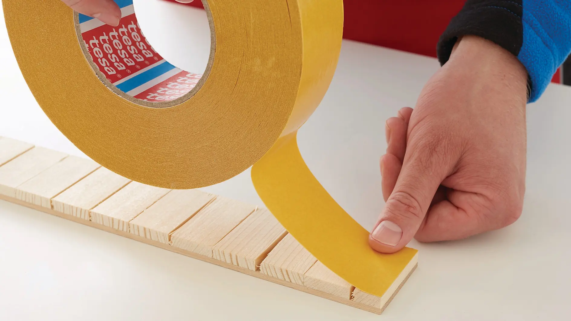 hand holding yellow adhesive tape, applying it to wooden slats on a white surface in an indoor setting