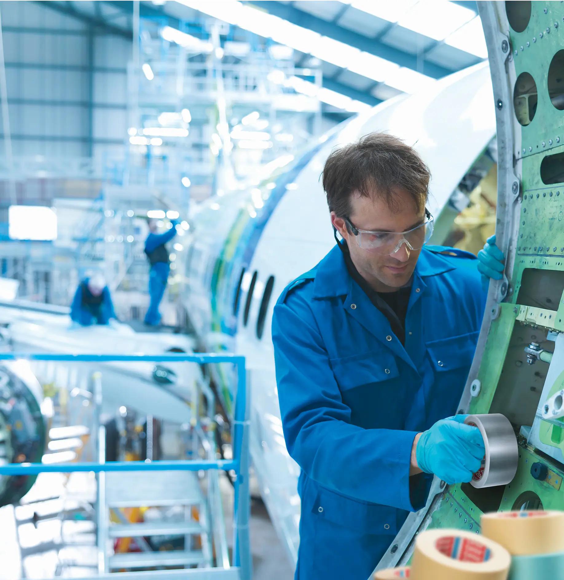 tesa tape being applied by a technician in blue coveralls inside an aircraft maintenance hangar with airplane parts visible in the background (This text has been generated by AI)